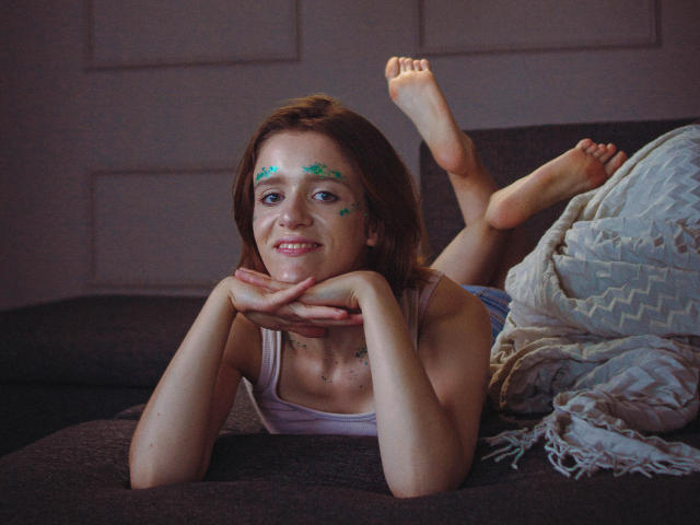 FrancescaL is lying on a couch, legs up, wearing a white tank top, green starface paint on her forehead, smiling, hands under her chin, bare feet in the air.