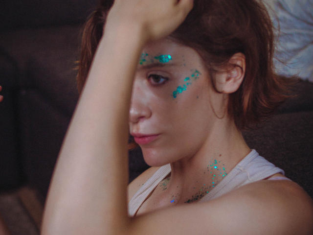 FrancescaL has sparkly blue glitter on her face and chest, brown hair, and a white tank top, looking sideways with her arm over her head in a selfie.