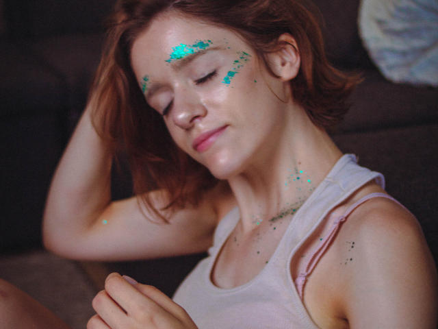 FrancescaL with red hair, white tank top, green glitter on face, eyes closed, hand in hair, soft smile, slight freckles, sitting on dark couch, relaxed, gentle expression.