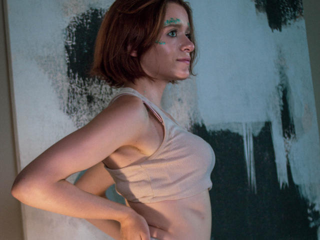 FrancescaL with red hair, green paint on her forehead, wearing a beige tank top, side profile, hands on hips, showing her tits and a slim, pale body.