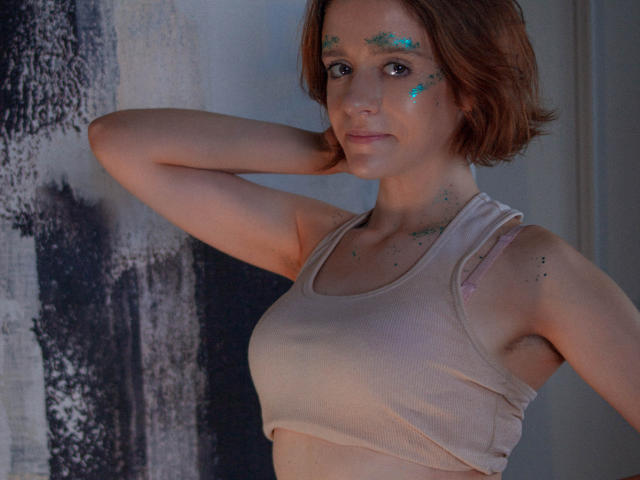 FrancescaL with short brown hair, wearing a beige sports bra, has glitter on her face and chest, arms raised, smiling subtly. Boobs visible, no pants, indoors.