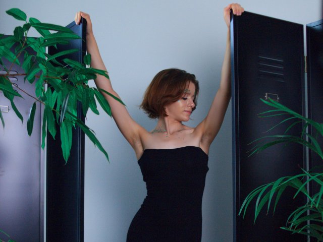FrancescaL with arms raised, wearing a black strapless dress, standing between black lockers, green plant leaves in front and to the side. She has short brown hair and a necklace.