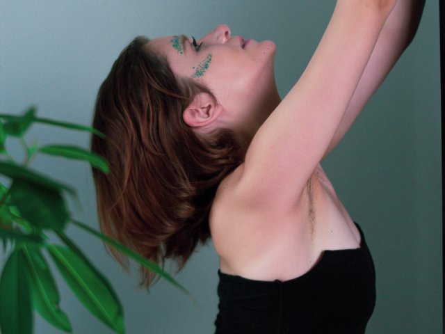 FrancescaL with green eye makeup, brown bob, black strapless top, arms raised, head tilted back, green plant in left. Her tits are small, arms are exposed.
