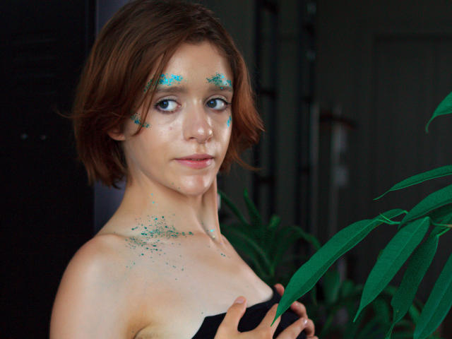 FrancesaL has short brown hair, green glitter on her face and chest, and is holding a black strap. She's standing next to a green plant with her tits exposed.