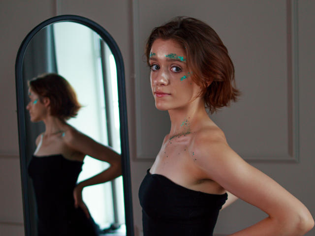FrancescaL has short brown hair, blue eyes, and green paint on her face, wearing a black strapless dress, standing in front of a mirror, showing her reflection.