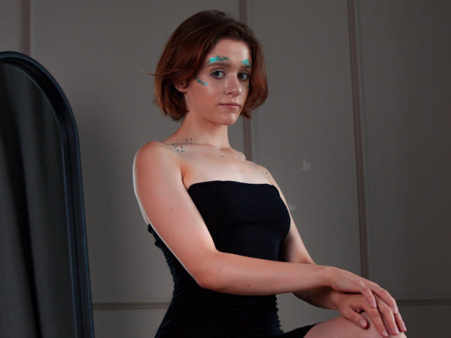 FrancescaL has short brown hair, blue face paint, and a black strapless dress, sitting with hands on knees, showing small tattoos on her chest. Her tits are visible.