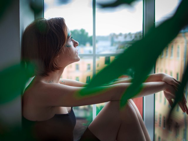 FrancescaL with short brown hair, black top, sitting by window, arms crossed, green plant leaves in foreground, looking outside, soft light, slight smile, calm expression, urban background.