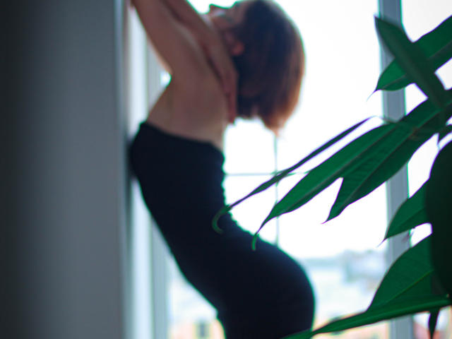 FrancescaL with short brown hair, topless, arms raised, wearing black pants, standing near a window, green plant in foreground, soft light, blurred background.