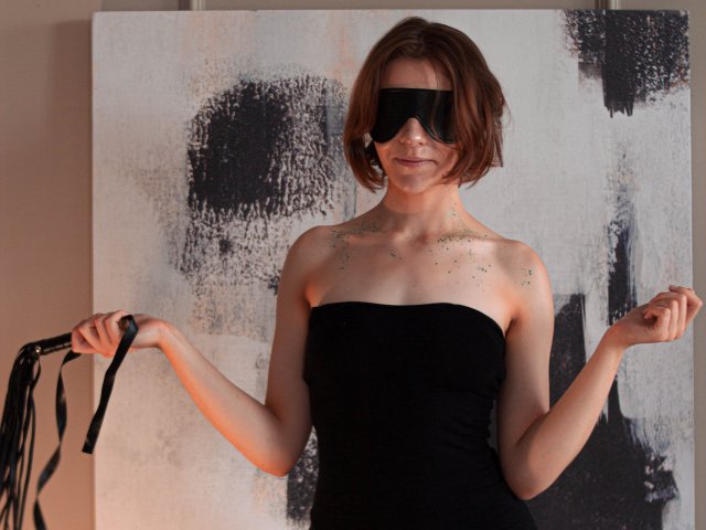 FrancescaL with short brown hair, black blindfold, strapless black dress, holding black leather flogger, arms raised, light green glitter on chest, standing in front of abstract black-and-white painting.
