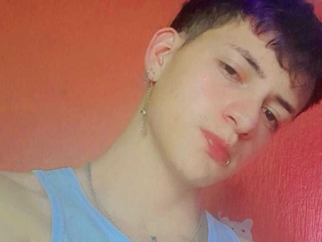 GabyThoms with short, dark hair, pink nose ring, and light blue shirt, wearing a chain necklace and earring, looking slightly to the side with a subtle pout.