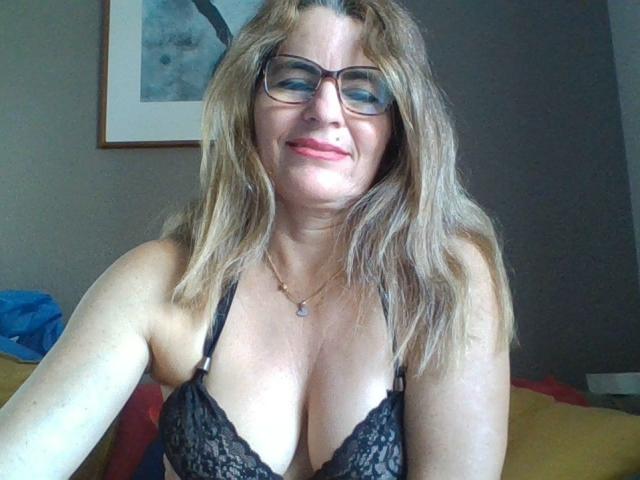 AliciaJoe with blonde hair, glasses, and black lace bra, showing off her big tits, smiling at the camera, in a bedroom with a painting on the wall.
