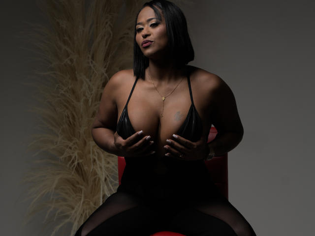 BeckyVega with dark skin, black bob, wearing a black halter top, covering her big, dark, round boobs with her hands, sitting on a red chair, looking confident.