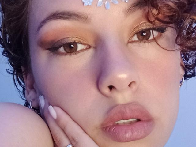 CristineBonnye's face with curly brown hair, brown eyes, orange eyeshadow, white leaf headpiece, pink lips, hand with pink nails touching cheek, blue background, and soft light.