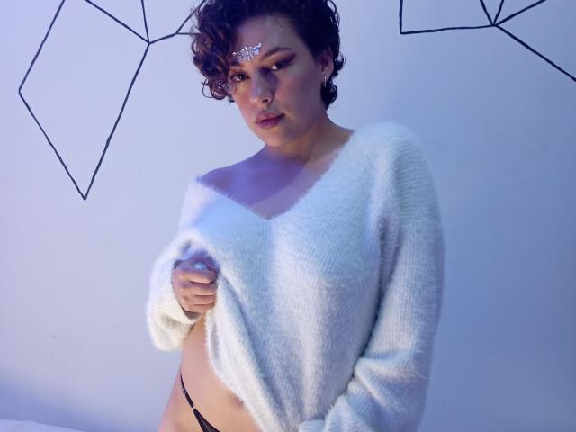 CristineBonnye with curly brown hair, wearing a white fuzzy sweater, holding it up to reveal black thong, has a jeweled forehead, and looks seductive with blue lighting.