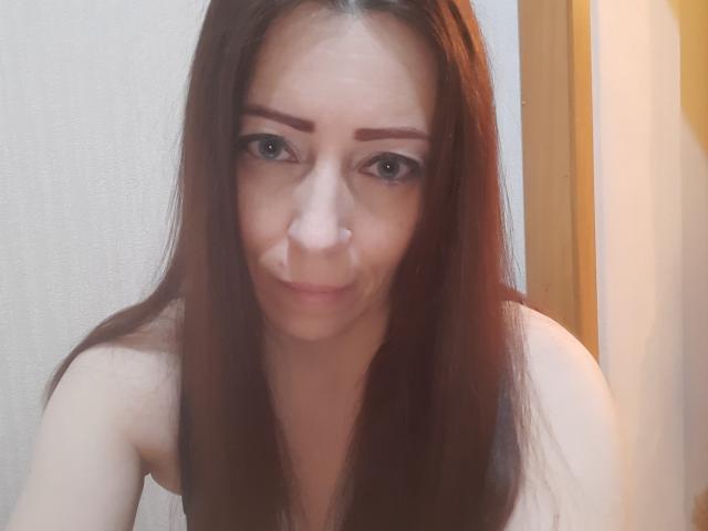 MaryanaLady with long brown hair, dark eyebrows, nose ring, and subtle smile, showing her tits, looking directly at the camera. Soft lighting, slight blur.