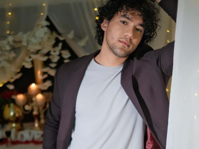 FreyrStyle69 with curly black hair, light brown skin, and a slight beard, wearing a dark purple jacket over a gray shirt, leaning against a white curtain.