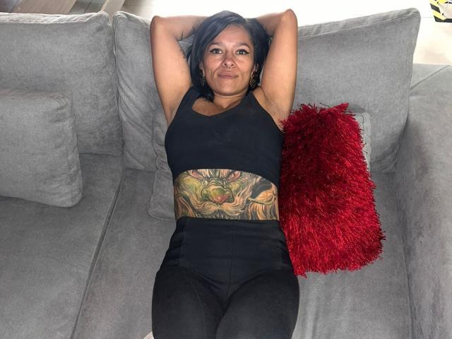 Amelie Blake, les bras derrière la tête, portant un débardeur de sport noir et un pantalon noir, avec un grand tatouage coloré sur le ventre, allongée sur un canapé gris avec un oreiller moelleux rouge, cheveux noirs, léger sourire..