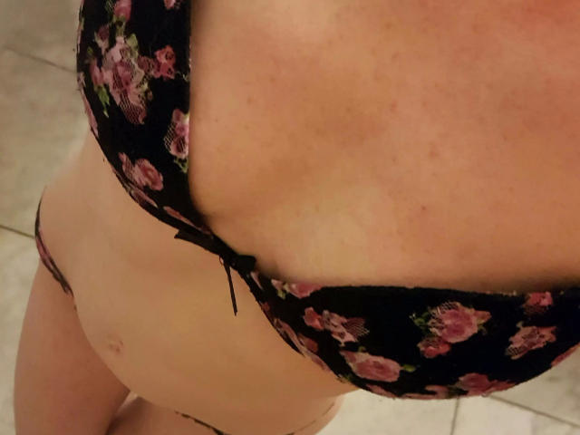 KellyBelly-hot's tits in a black floral bra, showing her pale skin and slight belly. She's wearing matching black floral panties, with a small black bow at the center.