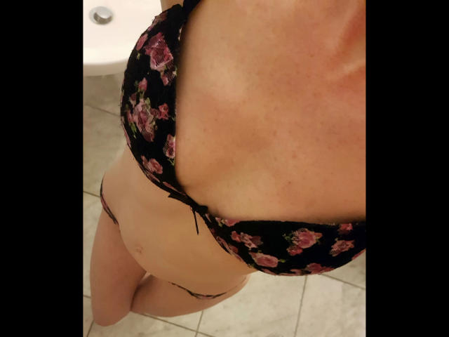 KellyBelly-hot's tits are in a black floral bra, showing tan lines, with matching panties, standing on tiled floor. Her light skin is slightly flushed.