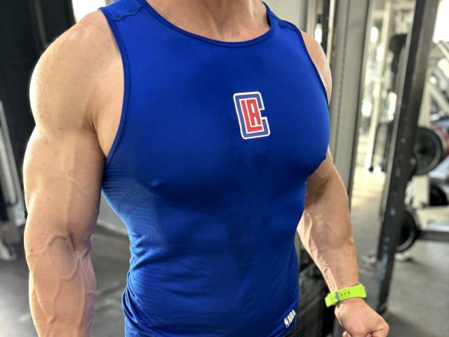 DestinyChris é um homem musculoso, vestindo uma camiseta azul que exibe seus braços e peito definidos. Ele está usando uma pulseira verde e tem um logotipo "C" na sua camiseta..
