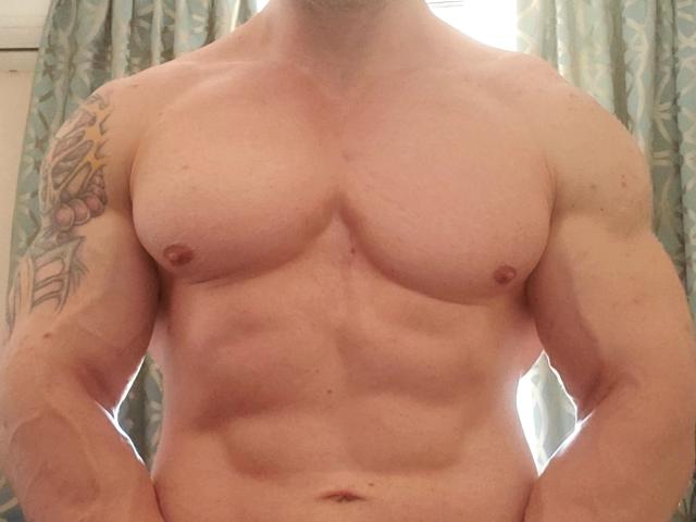 DestinyChris tiene un torso musculoso y sin camisa, con abdominales visibles, pezones pequeños y un tatuaje en el brazo izquierdo. Las manos de DestinyChris están en sus caderas, mostrando su pecho..