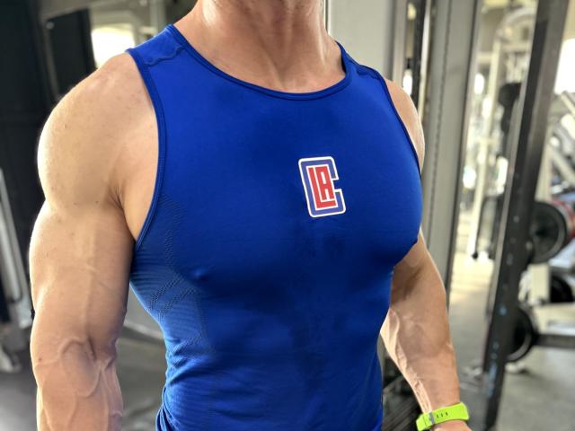 DestinyChris heeft een gespierd, getraind bovenlichaam met duidelijke biceps en draagt een strak, blauw shirt zonder mouwen, waardoor hun grote, zichtbare en stevige borst zichtbaar is. Ze dragen een groene polsband..