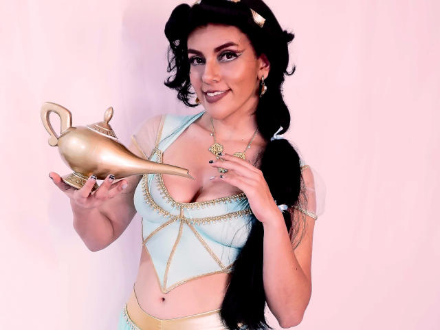 ZoeeMar with black pigtails, wearing a gold headpiece, blue and gold top, holding a gold genie lamp, smiling, showing her tits, pink background.