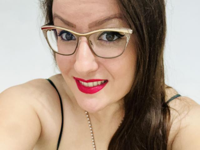 BadAngel with red lipstick, glasses, and a pearl necklace, showing off her tits in a black spaghetti strap top, smiling at the camera.