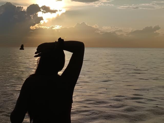 Silhouette de Katherin, d'origine latine, bras levé, debout dans une eau calme au coucher du soleil. La lumière du soleil est derrière elle, le ciel est nuageux, il y a un petit rocher dans l'eau et de légères vagues. Elle a de longs cheveux et on voit son poignet..