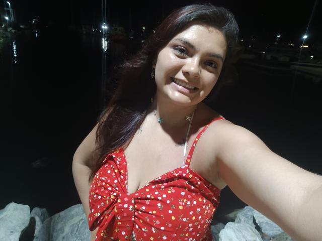 KatherinLatin with medium tits, smiling, wearing a red dress with small yellow patterns, dark hair, night, rock in foreground, visible necklace, holding phone, dimly lit, casual selfie.