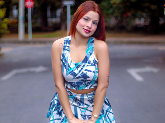 ShantalSummer with red hair, wearing a blue and white geometric crop top and skirt, sitting on a street, hands on knees, smiling, showing small tits, tattoo on right arm.