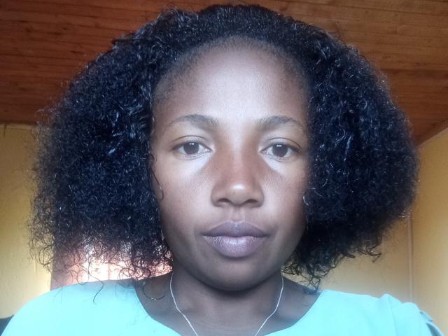 MoinnaJolie tem a pele marrom escura, cabelo preto encaracolado e lábios cheios com uma expressão neutra, vestindo uma camisa verde clara e um colar fino..