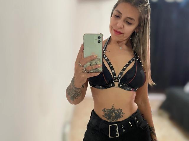 GuadaFeliz-hot with blonde hair, black leather harness, and tattoos, holding a phone, showing her tits and belly tattoo, wearing black pants, standing in a room.