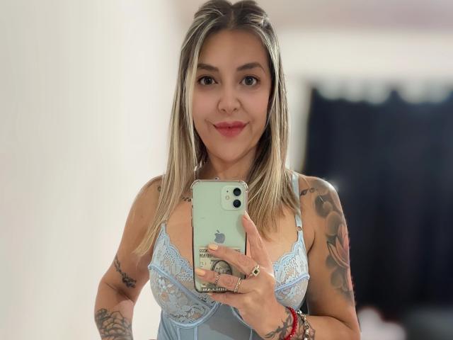GuadaFeliz-hot with blonde hair, blue lace bra, tattoos, holding iPhone, smiling at mirror, showing tits, wearing red bracelets, in soft lighting.