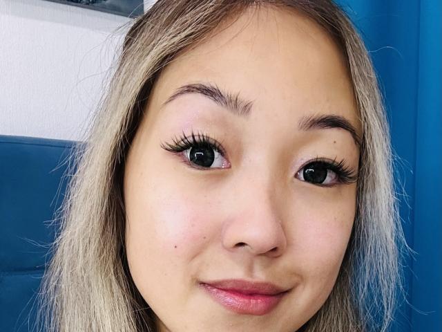 RenyLime's face, showing big dark eyes, long eyelashes, pink lips, and blonde hair, with a slight smile, against a blue background. Her tits aren't visible in this shot.