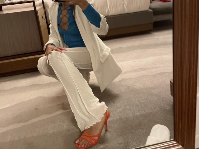 MissFeetParis with a blue top showing her tits through a cutout, white pants, and orange heels, squatting in front of a mirror, wearing a white coat.