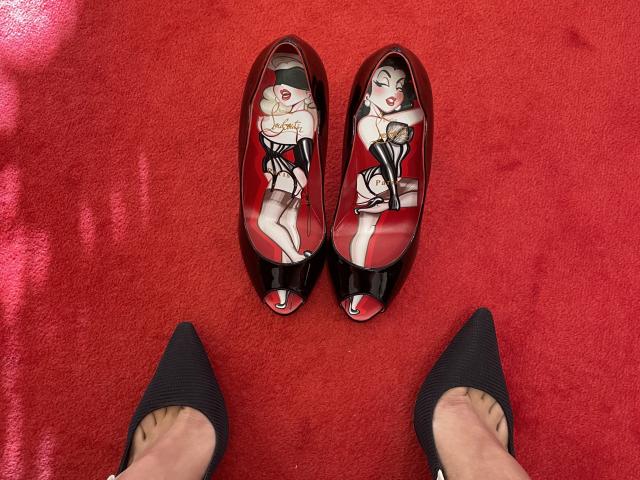 MissFeetParis' red, black, and white high-heeled shoes with a pin-up girl design, next to her black pointed-toe heels, on a red carpet. Her toes are visible.