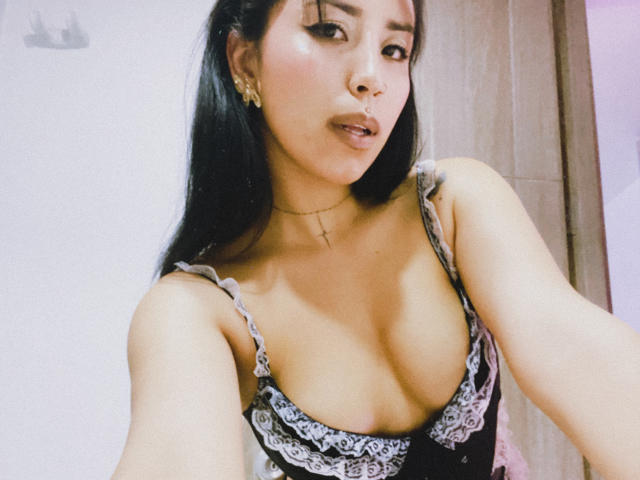 ElizabethMacy with long black hair, wearing a black and white lace-trimmed top, showing her big tits, looking at the camera, with a gold cross necklace and earrings.