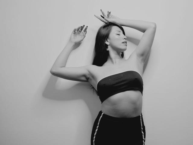 ElizabethMacy with her arms raised, wearing a strapless top and black pants, showing her toned abs and small tits, with long dark hair and closed eyes. Black-and-white photo.