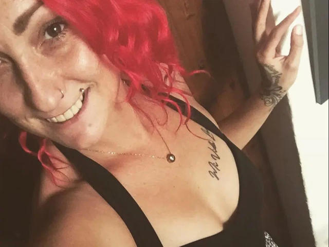 Wild-Vanilla-hot with red curly hair, smiling, showing her tits in a black tank top, tattoo on chest, nose ring, and tattoo on right arm, making a peace sign.