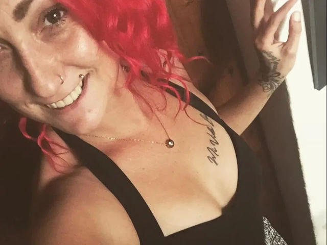 Wild-Vanilla-hot with red curly hair, nose ring, smiling, black tank top, visible tattoo on chest, holding phone, small breasts, necklace with pendant, tattoo on right hand.