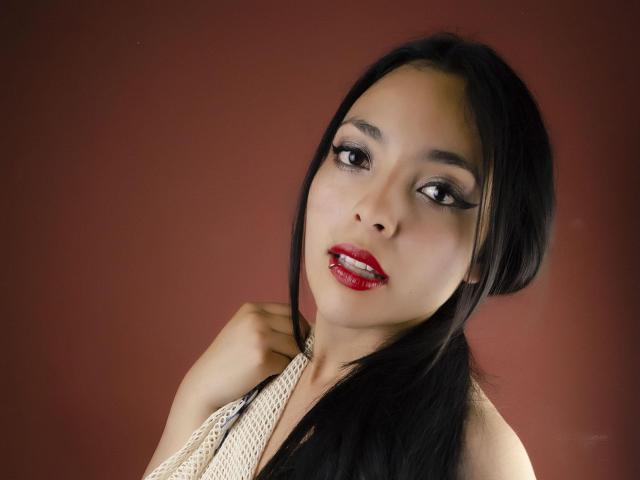 SakuraHarper ha i capelli neri, il rossetto rosso e uno sguardo seducente con un trucco per gli occhi marcato. Indossa un top a fascia, con una mano sulla spalla, e guarda leggermente di lato..