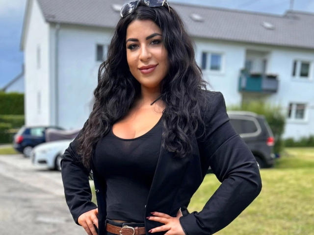 LadyAyse-hot with wavy black hair, wearing a black top and jacket, brown belt, and sunglasses on head, hands on hips, smiling, standing in front of a white house.