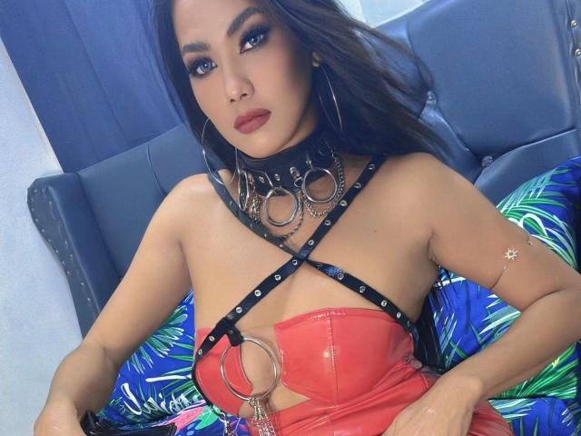 AddictiveMiss4u-hot with dark hair, wearing a red leather top, black harness with metal rings, and choker, sitting on a blue couch, looking at the camera with red lipstick.