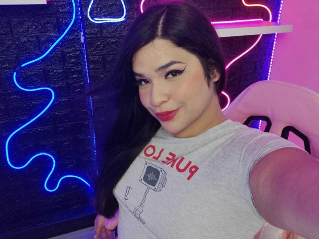 AntonelaPark with long black hair, red lipstick, and eyeliner, wearing a gray T-shirt with a camera print, smiling at the camera, neon lights in the background.