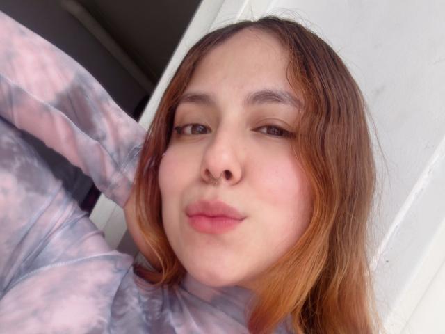 AmberlStha has light skin, brown eyes, and pinkish lips. She's wearing a pink and gray tie-dye hoodie with her brown hair falling to the sides.