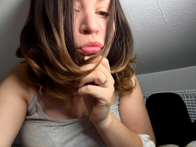 AnnabellkaSecret with pouty pink lips, brown hair with blonde highlights, grey tank top, hand touching chin, ring on finger, slightly messy hair, white ceiling, soft lighting.