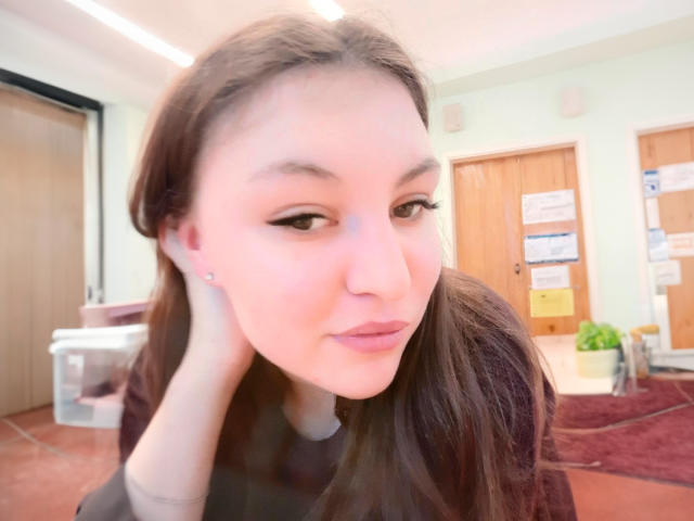 AnnabellkaSecret with long brown hair, light skin, and black eyeliner, looking slightly to the side, holding her head with a small nose piercing, in a softly lit room.