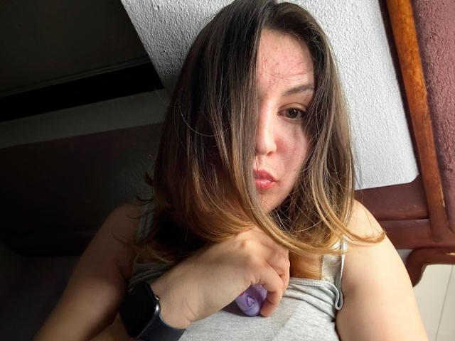 AnnabellkaSecret with brown hair, pouty lips, and a purple vibrator in her hand, wearing a gray top, looking at the camera, wearing a black wristwatch.