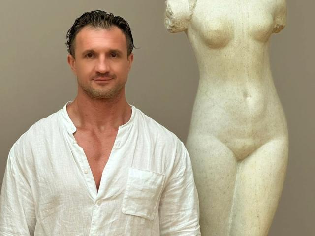 DestinyChris, con el pelo oscuro y mojado, vestida con una camisa blanca abotonada, está de pie junto a una estatua de mármol blanco con senos, trasero y vulva..